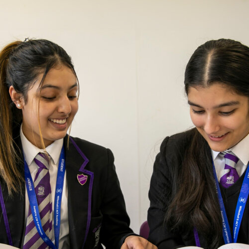 Two teens smiling reading their text books in school. They wear pins of Community Ambassador and Equality Ambassador.
