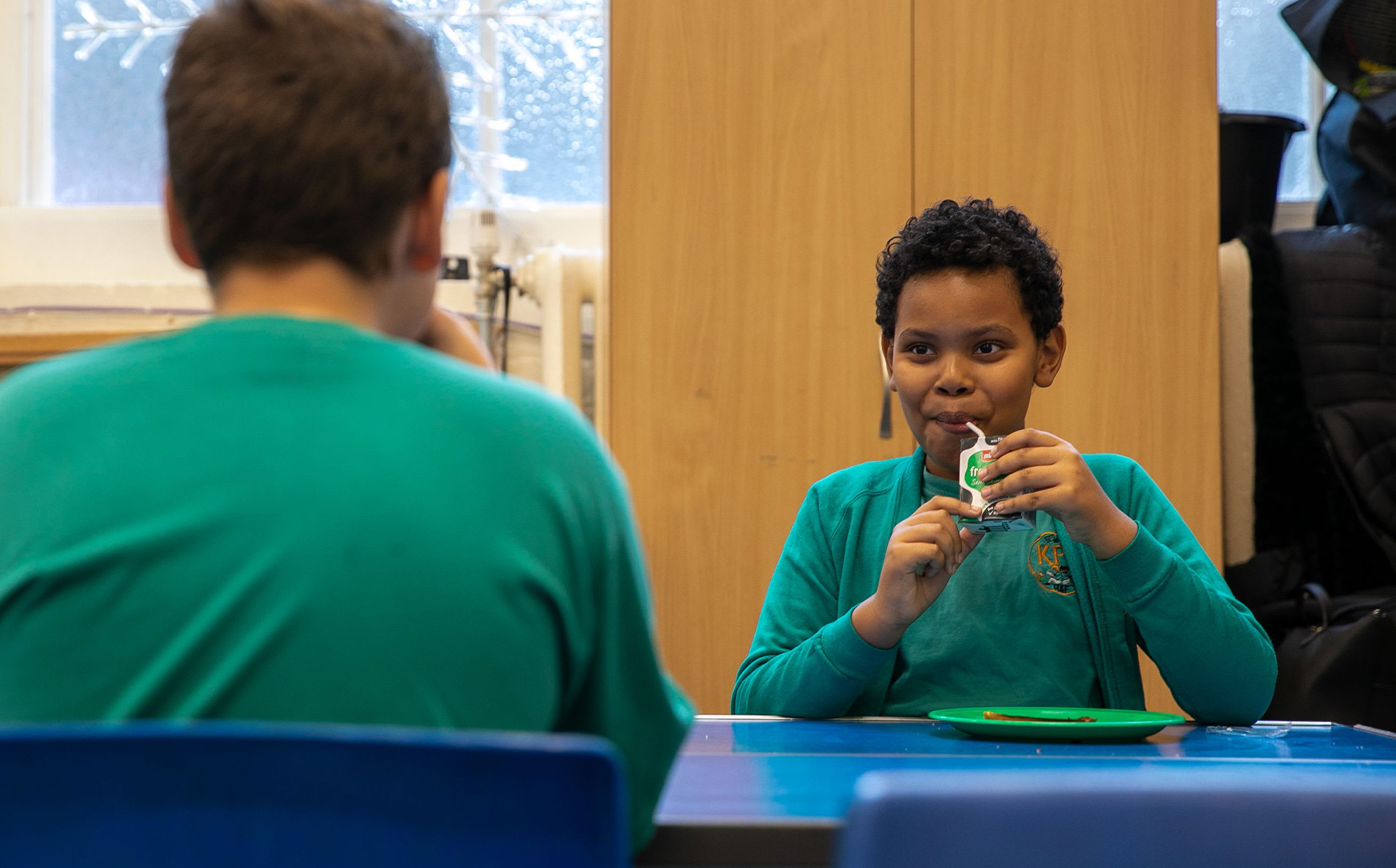 A boy smiling to his friend whilst drinking a carton of milk.