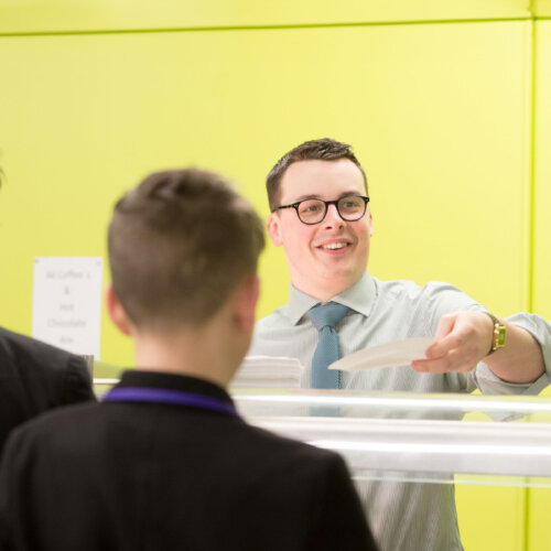 An adult serving breakfast in a yellow school cafeteria to teens in school uniform.