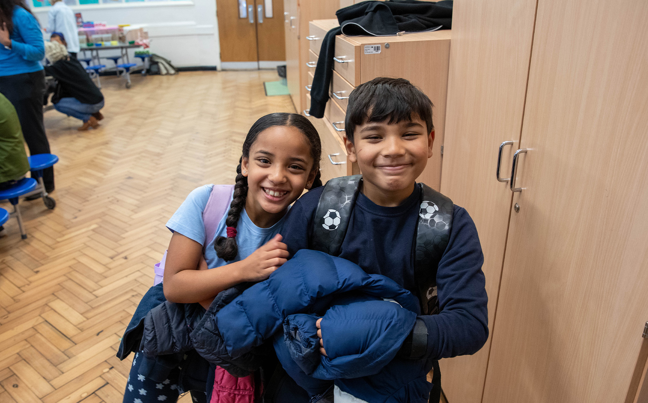 A photo of two childen smiling at the camera at school. The background is a table full of breakfast cereal.