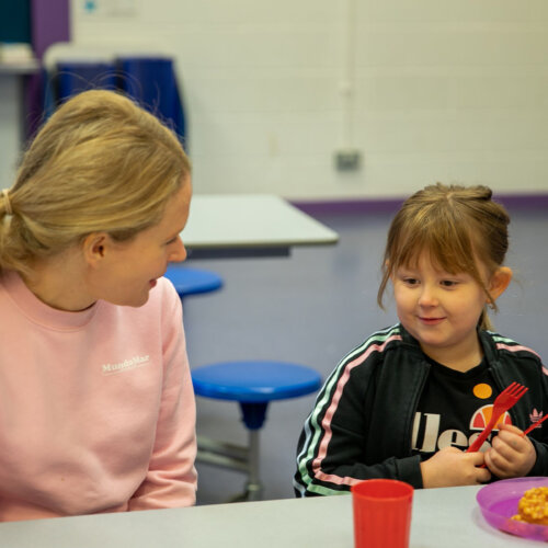 A photo of a young girl enjoying beans on toast at school and chatting to an adult seated next to her.
