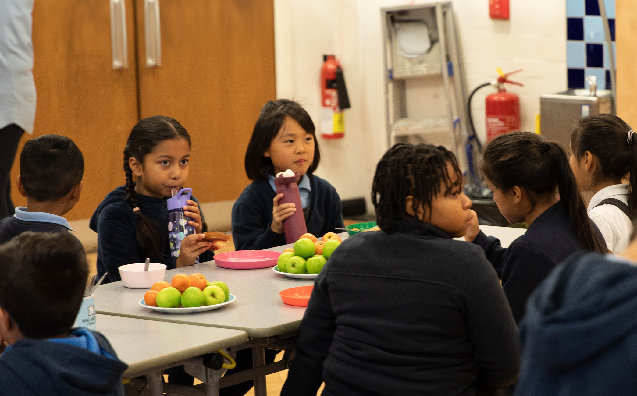A group of 7 children sitting on a table filled with fruit. They are enjoying toast and water.
