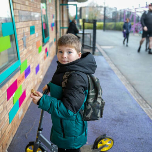Photo of boy in blue/black coat and backpack on a scooter looking at camera outside school.
