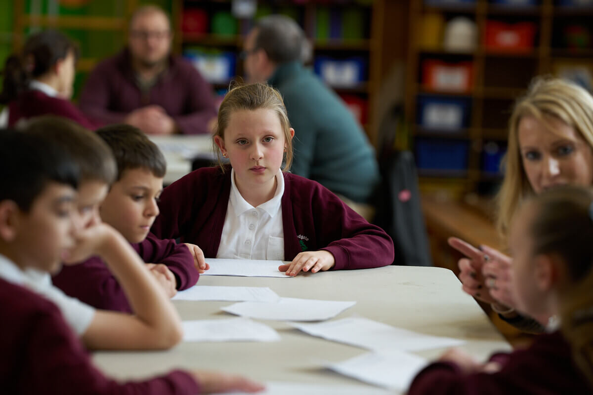 Child sitting at a table, looking at worksheets and engaging with classmates in a school setting