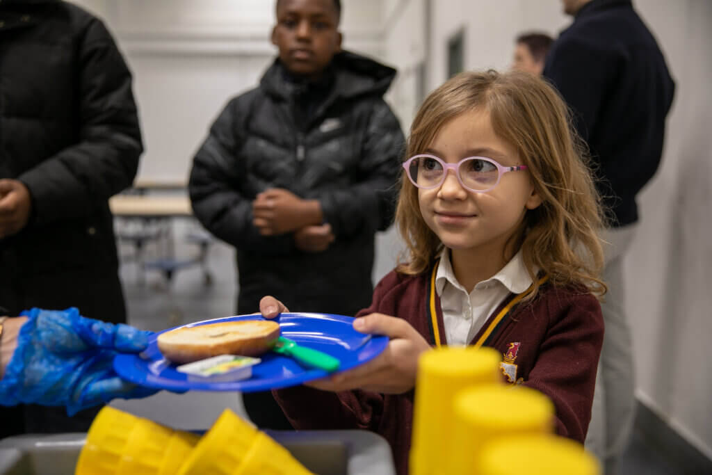 Image of a child being served a toasted bagel and spread on a plastic plate at breakfast club.