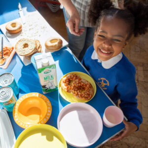 An image of a young child ages child in uniform with a big smile standing behind a table that had a spread of breakfast food including beans on toast, milk and bagels.