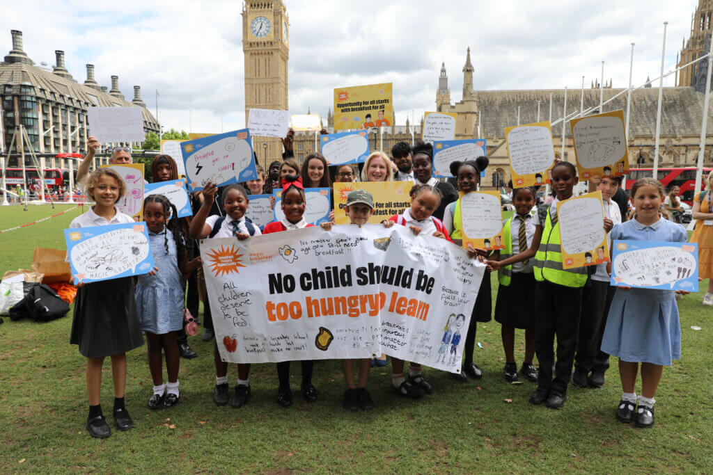 MP, Emma Lewell, stands with a large crowd of school children in front of Big Ben and the Houses of Parliament. They are holding a banner in front of them which reads, "No Child Should Be Too Hungry To Learn" and lots of the children are holding up placards with messages on the importance of school breakfasts.