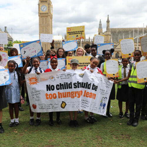 MP, Emma Lewell, stands with a large crowd of school children in front of Big Ben and the Houses of Parliament. They are holding a banner in front of them which reads, "No Child Should Be Too Hungry To Learn" and lots of the children are holding up placards with messages on the importance of school breakfasts.
