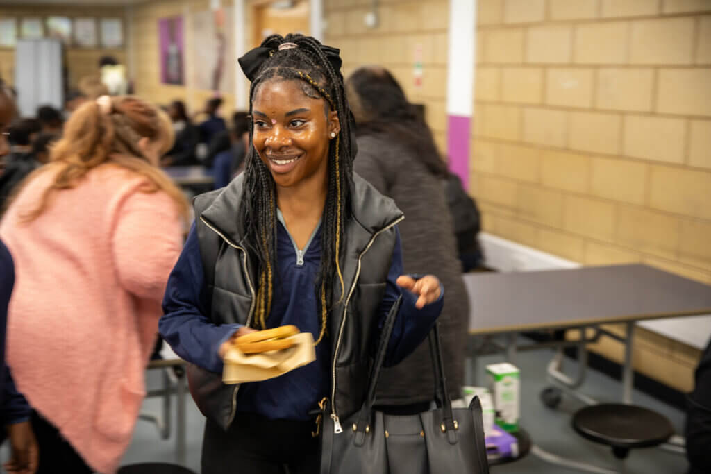 A secondary student walks away from the school breakfast bar, smiling and holding their freshly toasted bagel