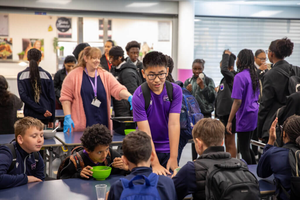 A secondary school student stands over a table full of their friends, talking as they enjoy their school breakfast