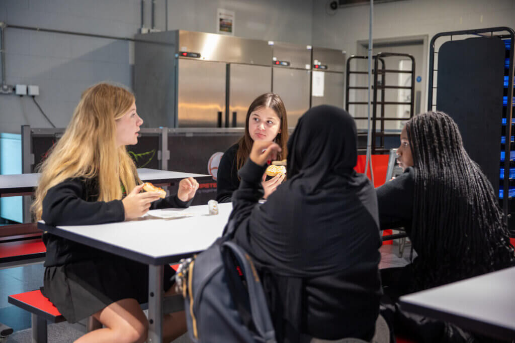 A group of secondary students sit in the canteen, talking and eating breakfast together before the school day begins