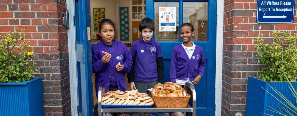 Three primary students stand proudly behind a trolley of breakfast foods in their school playground