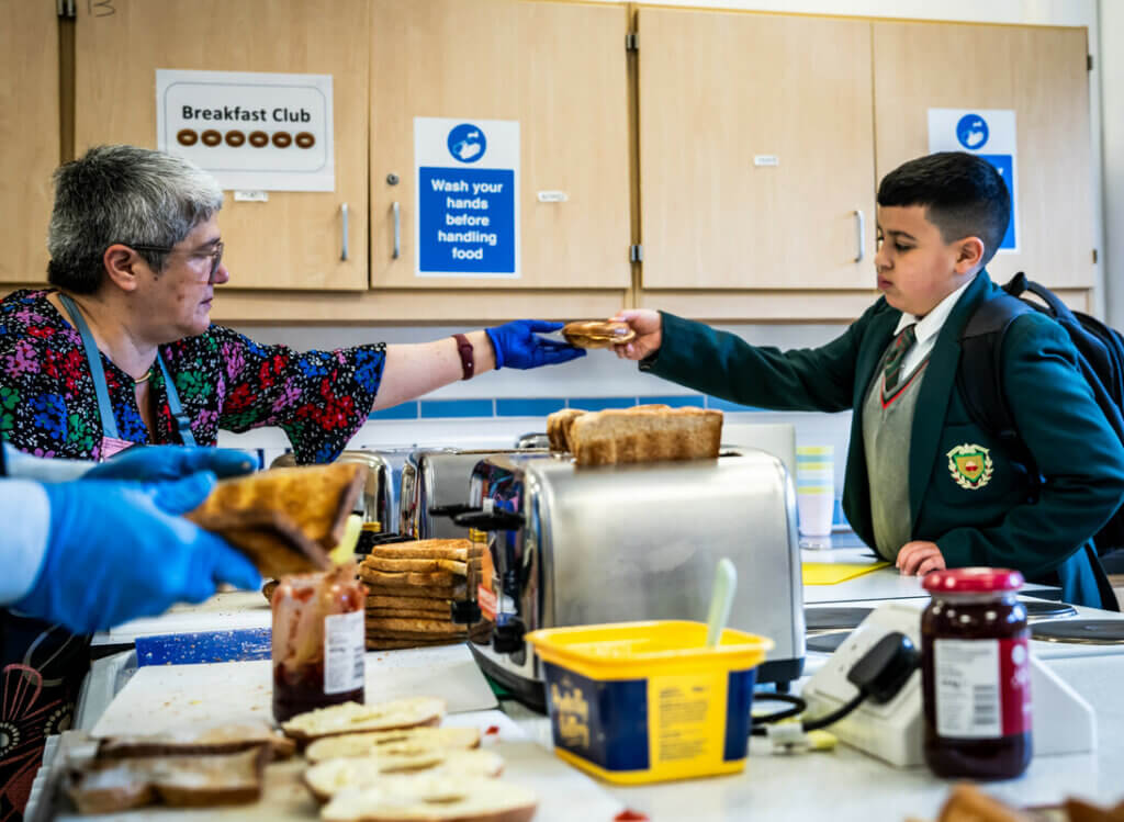 A secondary school teacher running the school breakfast club hands over a freshly toasted bagel to a student